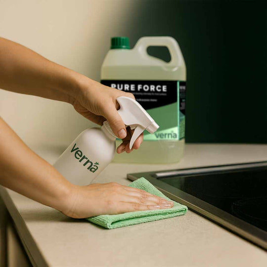 Person cleaning a countertop with a Verna spray bottle and green cloth, with a Pure Force bottle in the background.